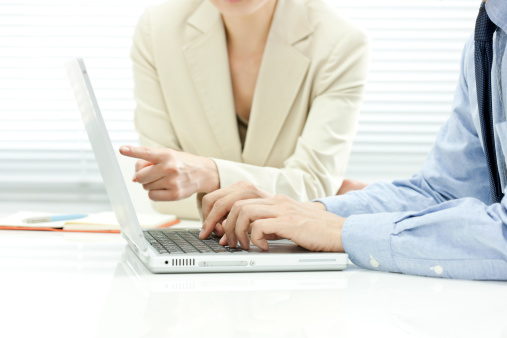 A businessman typing on a laptop that's on a table with a notebook, a pen, and business woman pointing at the laptop.